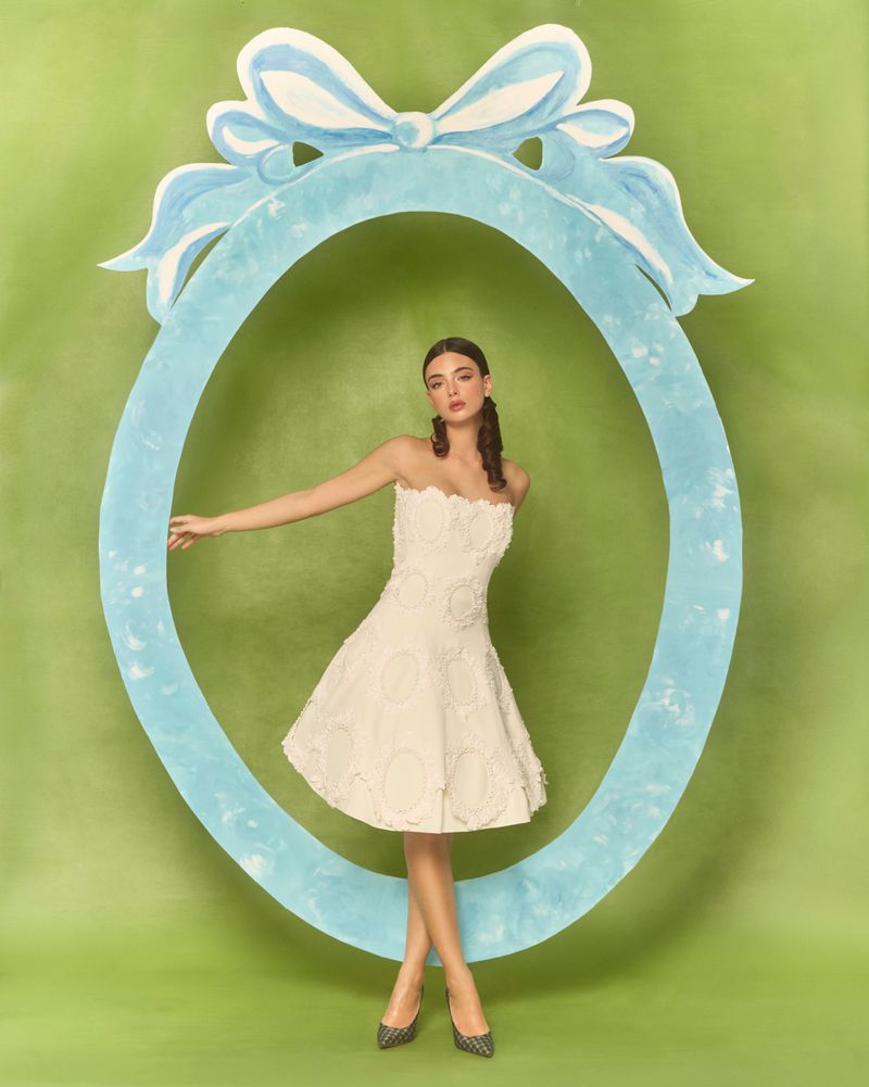 A woman in a short white dress poses within a light blue oval frame against a light green backdrop in a studio shot.