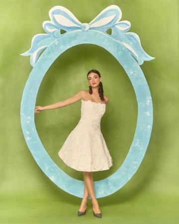 A woman in a short white dress poses within a light blue oval frame against a light green backdrop in a studio shot.