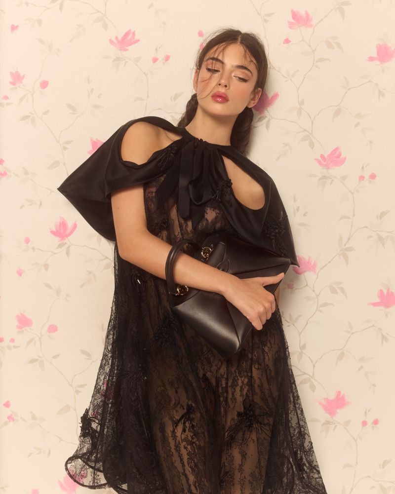 A woman in a sheer black lace gown poses elegantly against a pastel pink floral wallpaper.