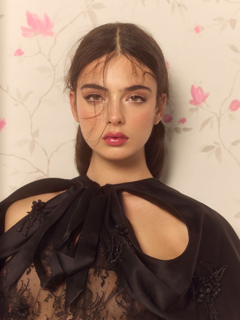 Headshot of a young fair-skinned woman with brown hair and dark eyes wearing a black lace top and cape, posing against a floral wallpaper.