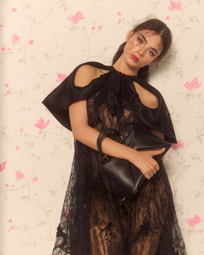 A young woman poses in a lacy black dress with a black handbag, against a light background patterned with pink flowers and green leaves.