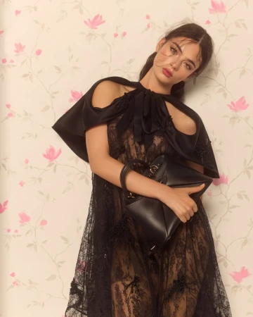 A young woman poses in a lacy black dress with a black handbag, against a light background patterned with pink flowers and green leaves.