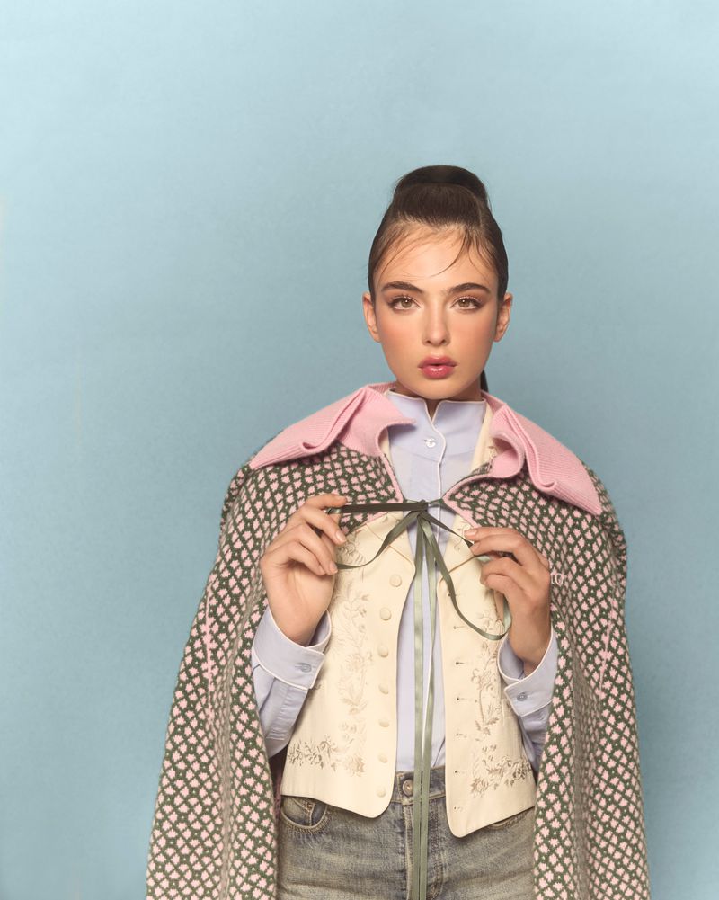 Portrait of a brunette woman in denim jeans, a patterned cape, and a pale vest, posing indoors against a light blue backdrop.
