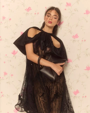 Deva Cassel wearing a black lace dress and holding a handbag against a floral wallpaper background.
