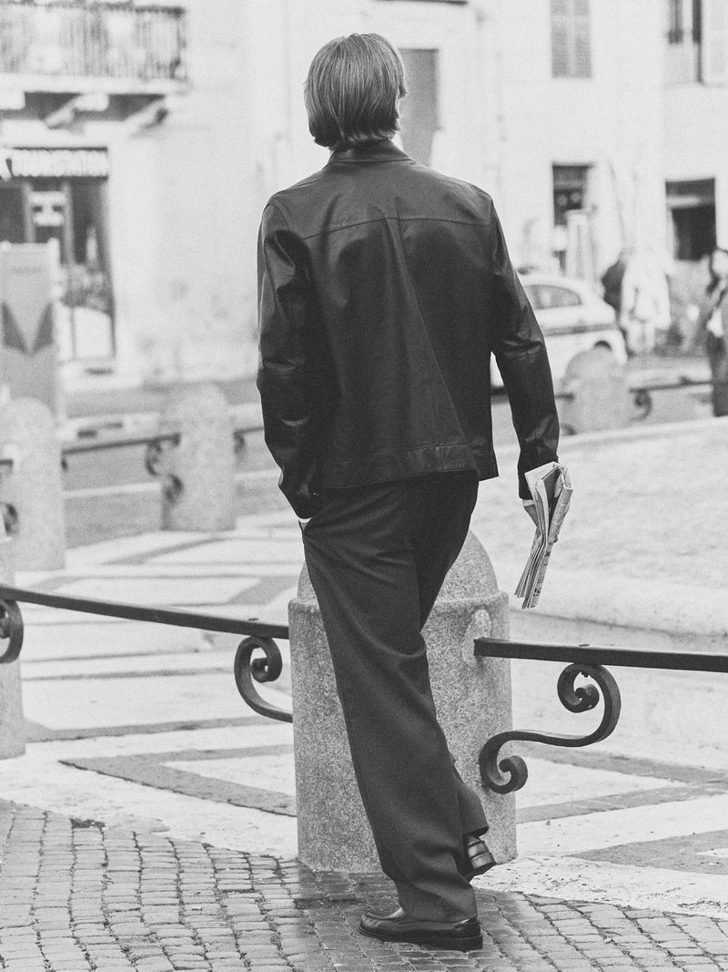 Man in black leather jacket and pants walking on a cobblestone street with a newspaper, leaning on a stone bollard in a European setting.