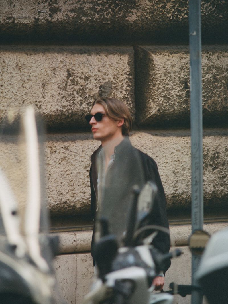 Young adult man with sunglasses stands near motorcycles on a sunny day in an urban environment with a stone wall background.