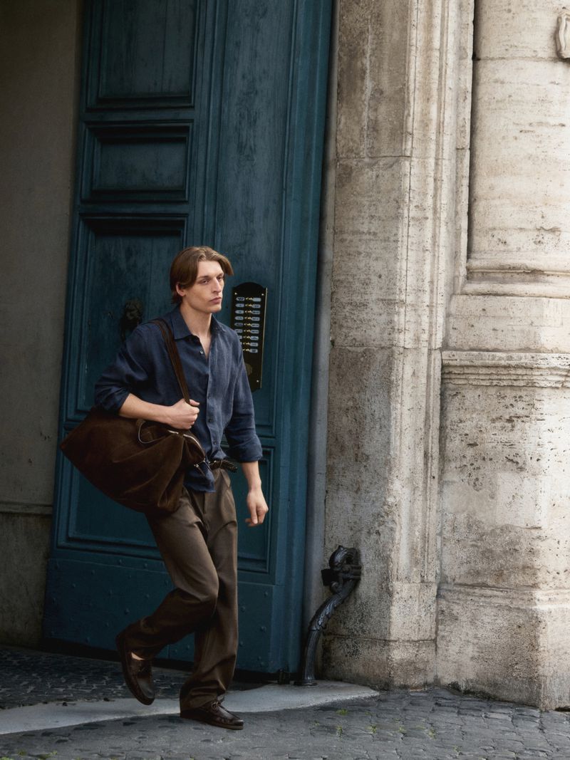 A young adult male carrying a brown duffel bag is walking out of a building with a large blue door and light stone facade in daylight.