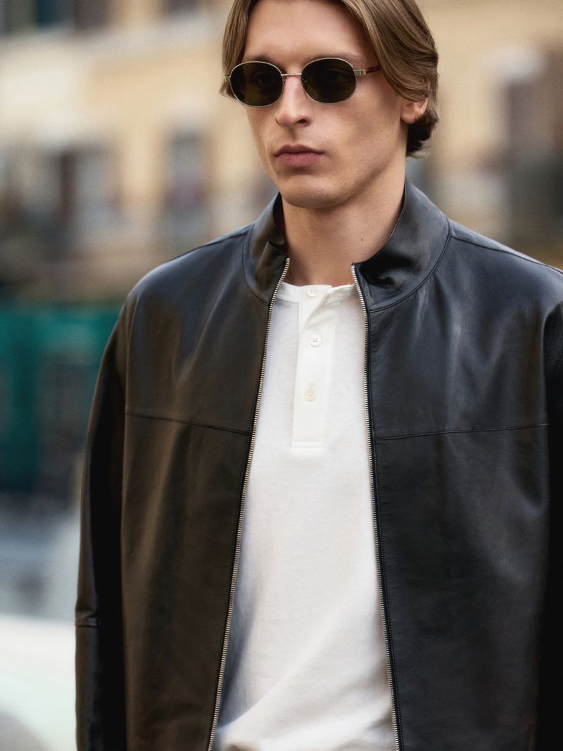 Man with blonde hair wearing dark oval sunglasses and a black leather bomber jacket over a white henley shirt, outdoor setting.