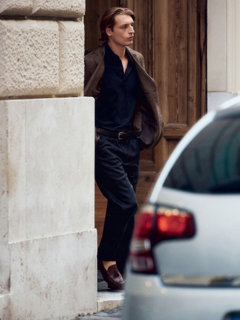 A fashionable man in a jacket and slacks exits a building toward a silver car, captured in natural outdoor light.