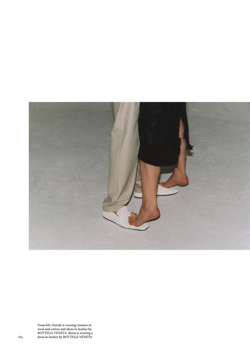 Two people are seen from the knees down, a man in beige pants and woven white loafers, and a woman in a black dress and loafers.