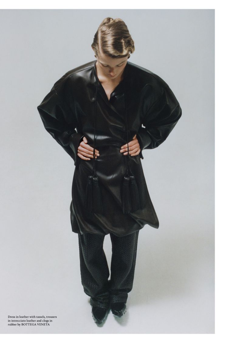 Fashion editorial shot of androgynous model wearing an oversized black leather tunic, wide trousers, and patent leather shoes in a studio.