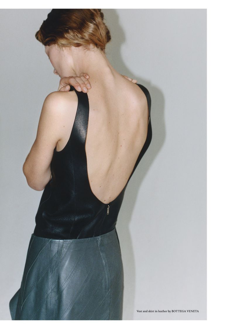 Fashion shot of a woman with red hair wearing a black leather top and grey skirt, posing in a studio with soft lighting.
