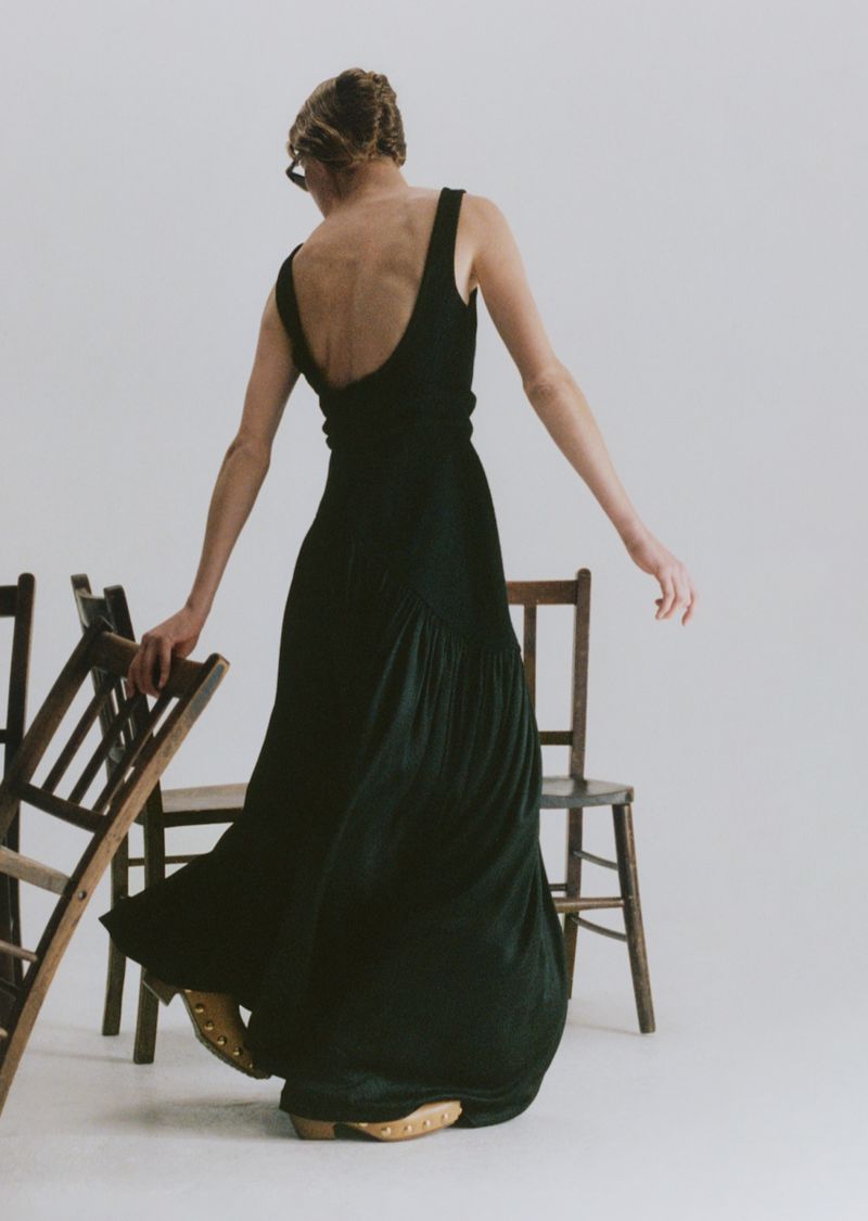 A woman with blonde hair stands in a photo studio, wearing a dark green dress and light-colored shoes, beside several wooden chairs.