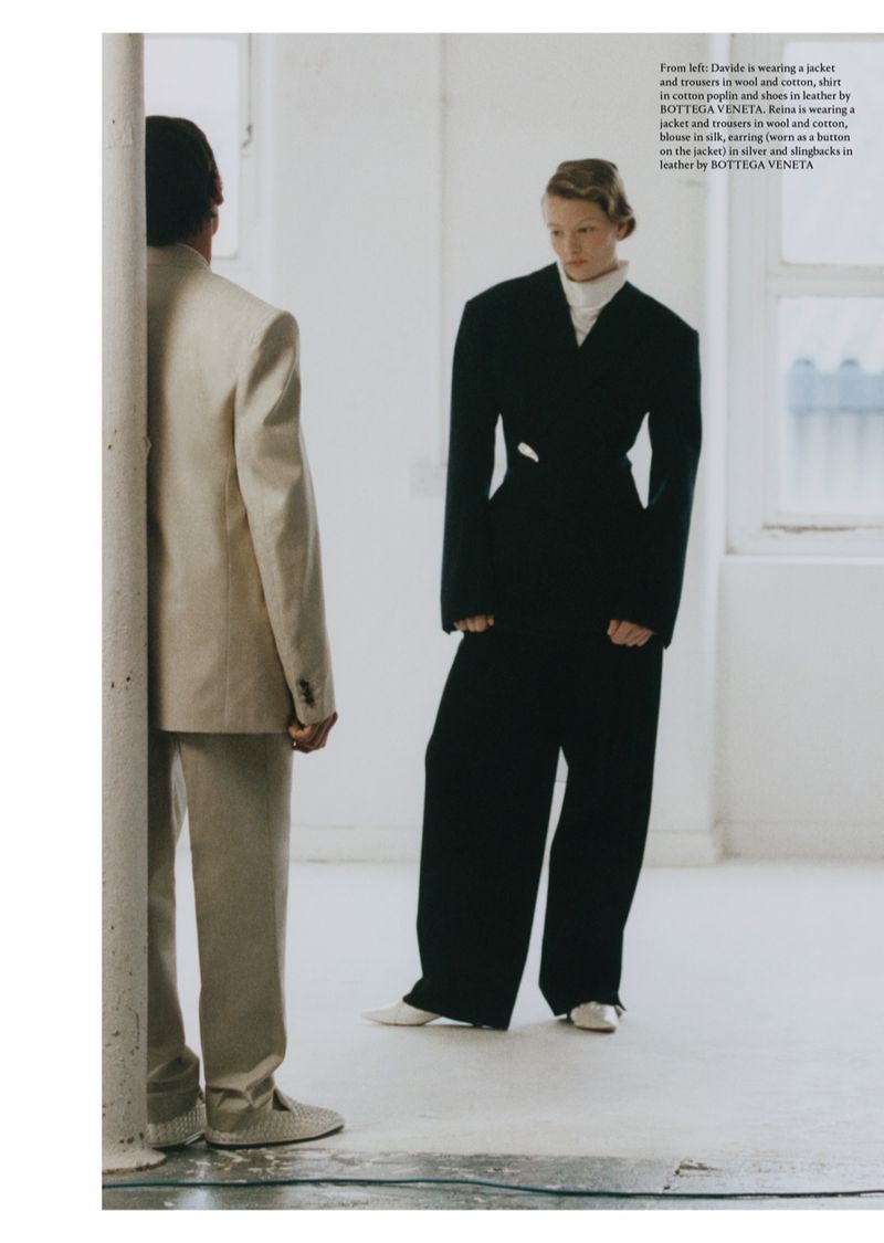 Two people in an indoor fashion shoot, one wearing a beige suit standing near a pillar and another wearing a black suit looking towards the right.