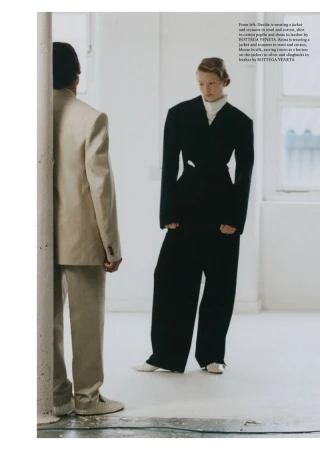 Two people in an indoor fashion shoot, one wearing a beige suit standing near a pillar and another wearing a black suit looking towards the right.