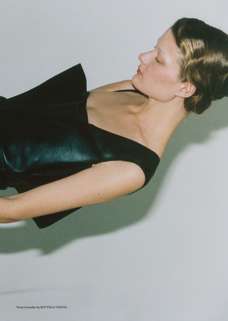 Fashion editorial shot of a model in a black leather dress against a pale background, captured in soft lighting with a shallow depth of field.