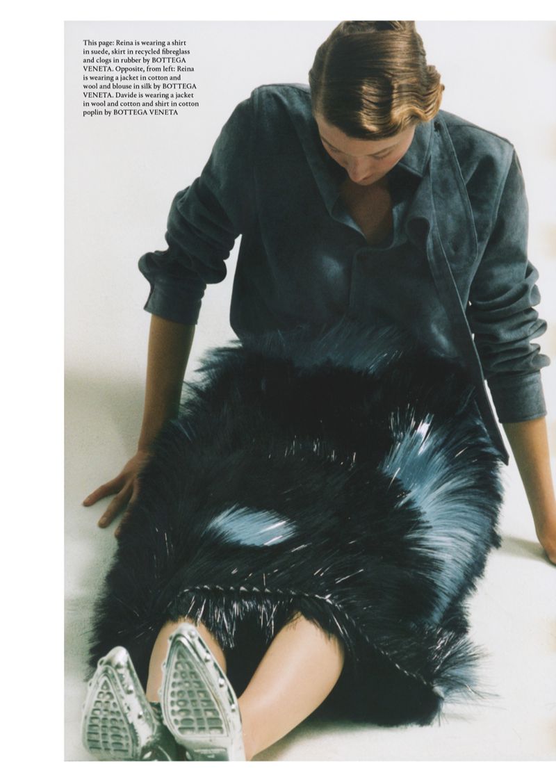 A person sits with legs extended, wearing a grey shirt, feather-like skirt, and silver shoes, in a fashion editorial shot.