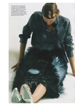 A person sits with legs extended, wearing a grey shirt, feather-like skirt, and silver shoes, in a fashion editorial shot.