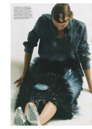 Model seated on white background wearing dark shirt and textured skirt with silver pointed shoes