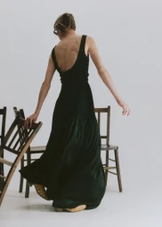Model in a black maxi dress touching wooden chairs against a plain white studio background