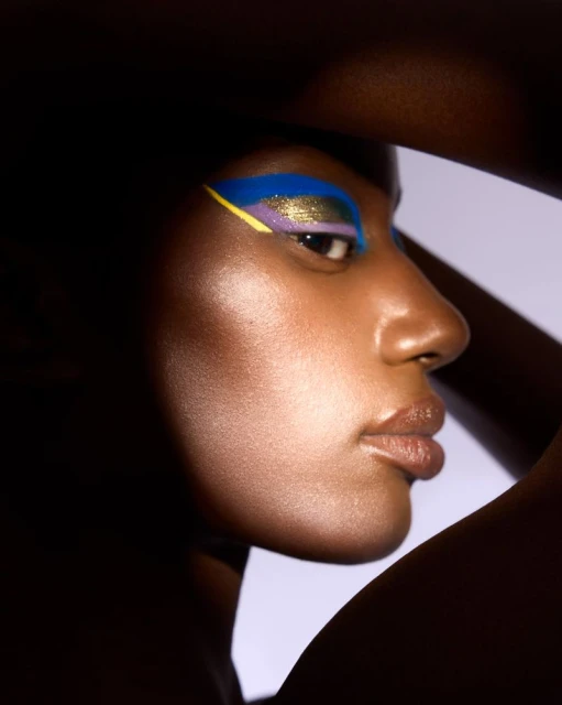 Close-up studio shot of an African American adult woman with colorful eye makeup, posing with a neutral facial expression against a light background.