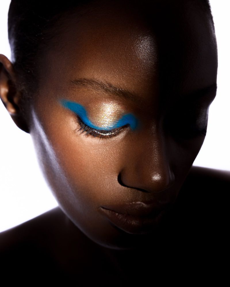 Close-up portrait of a Black woman featuring bold blue graphic eyeliner and metallic gold eyeshadow against a white background.