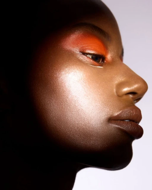 Close-up studio portrait of an African American woman with orange eyeshadow. Her skin has a dewy finish and the background is soft white.