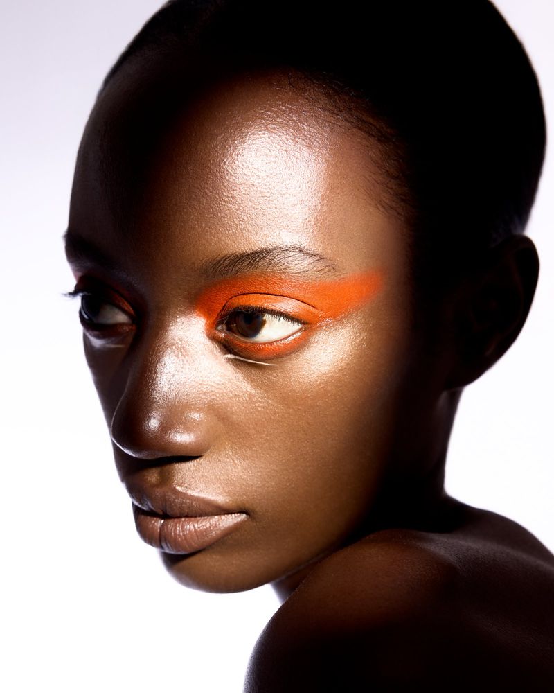 Close-up portrait of a woman with dark skin, orange eyeshadow, and glossy skin against a bright white background. Beauty and fashion.