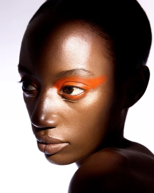 Close-up portrait of a woman with dark skin, orange eyeshadow, and glossy skin against a bright white background. Beauty and fashion.