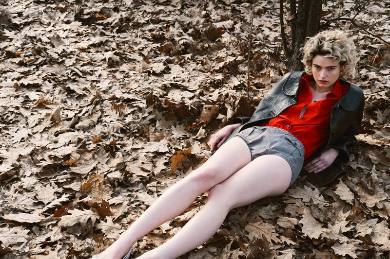 Woman in red top and grey shorts lies on a bed of autumn leaves in a woodland setting, posing for a fashion shot.