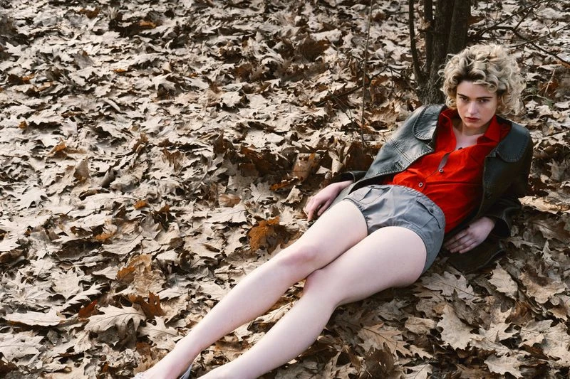 Woman in red top and grey shorts lies on a bed of autumn leaves in a woodland setting, posing for a fashion shot.