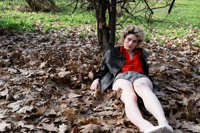 A young adult woman with curly hair rests in a pile of leaves next to a tree in an outdoor setting, wearing a red shirt and black jacket.