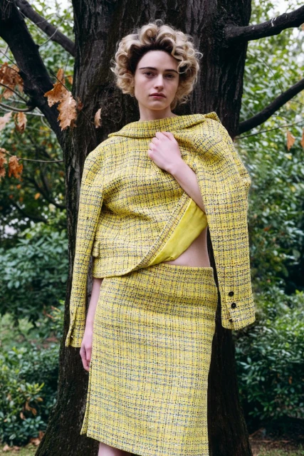 Woman poses in a yellow tweed skirt suit in an outdoor setting near a tree. The jacket is styled open, showcasing a matching yellow top.