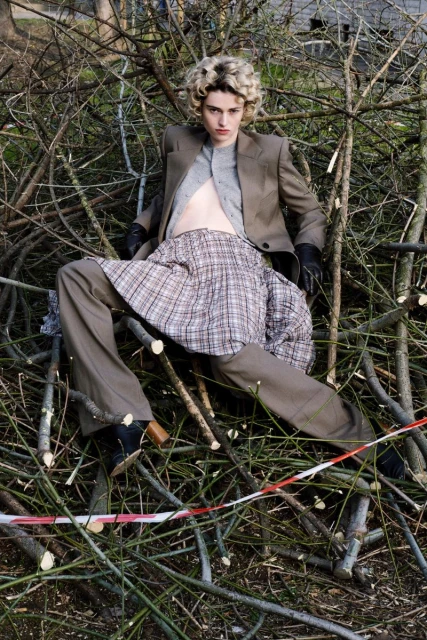 Fashionable young adult in couture outfit poses in a pile of tree branches in an outdoor setting under natural daylight.
