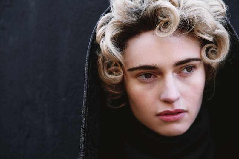 Close-up portrait of a woman with curly blonde hair and dark hooded outerwear against a black backdrop.
