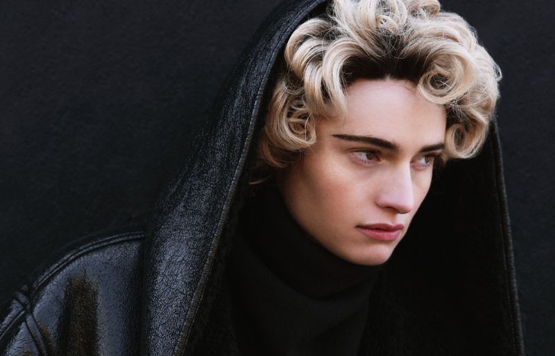 Close-up portrait of a person with blonde curly hair wearing a black turtleneck and a black hooded jacket against a dark background.