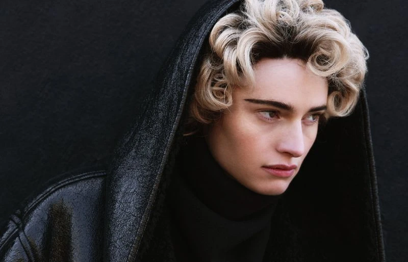 Close-up portrait of a person with blonde curly hair wearing a black turtleneck and a black hooded jacket against a dark background.