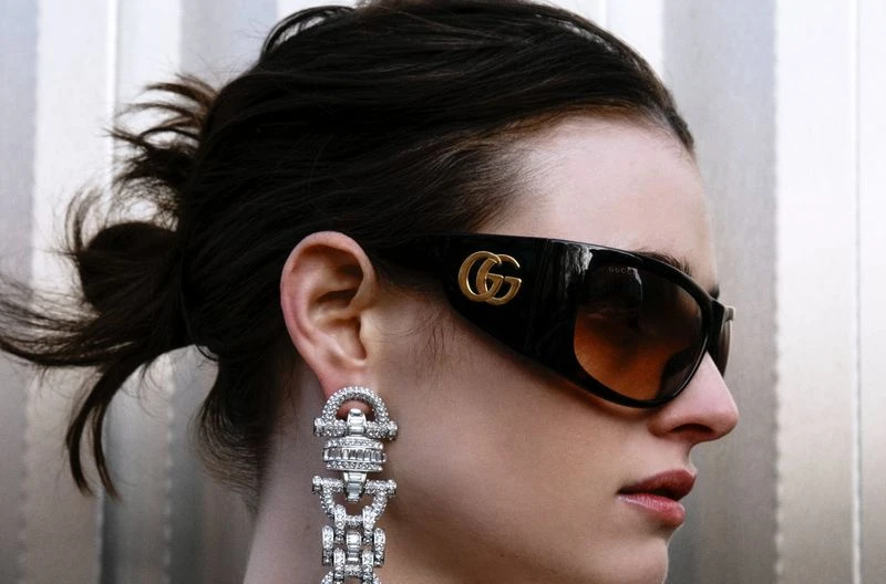Adult female in a medium close-up shot. She wears black sunglasses with a gold logo and large, glittering dangle earrings.