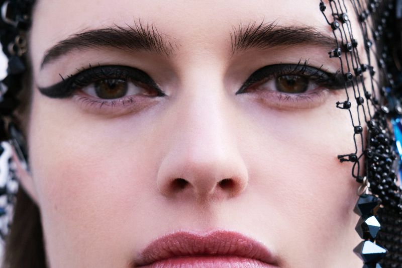 Close-up shot of a woman's face with striking black eyeliner and subtle lip color, accentuated by beaded headwear.