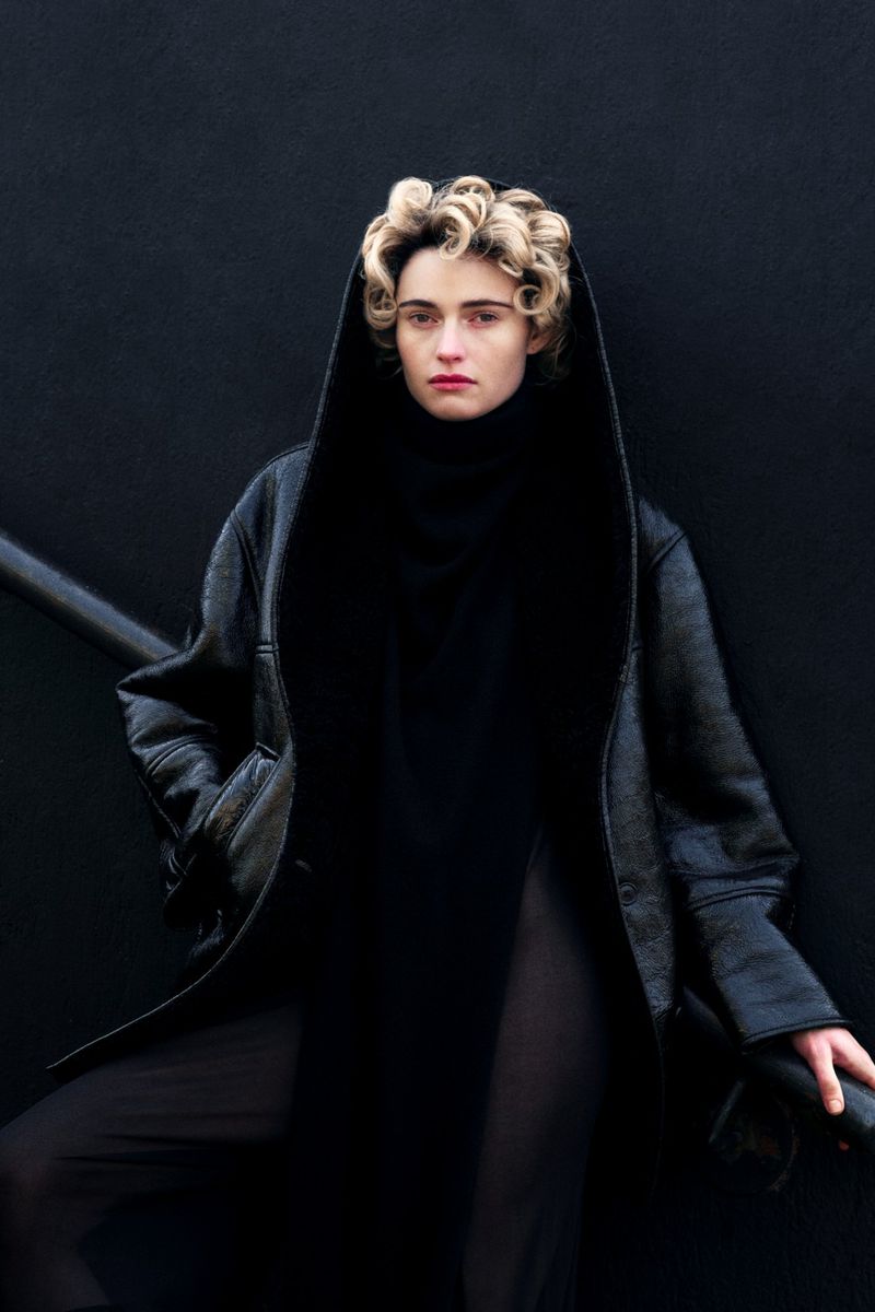 A woman with blonde curly hair and a serious expression poses in a black hooded coat and turtleneck against a black background.