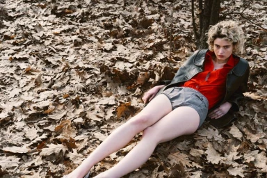 A blond woman lying amidst a thick layer of dry autumn leaves wearing a dark jacket, red shirt, and grey shorts.