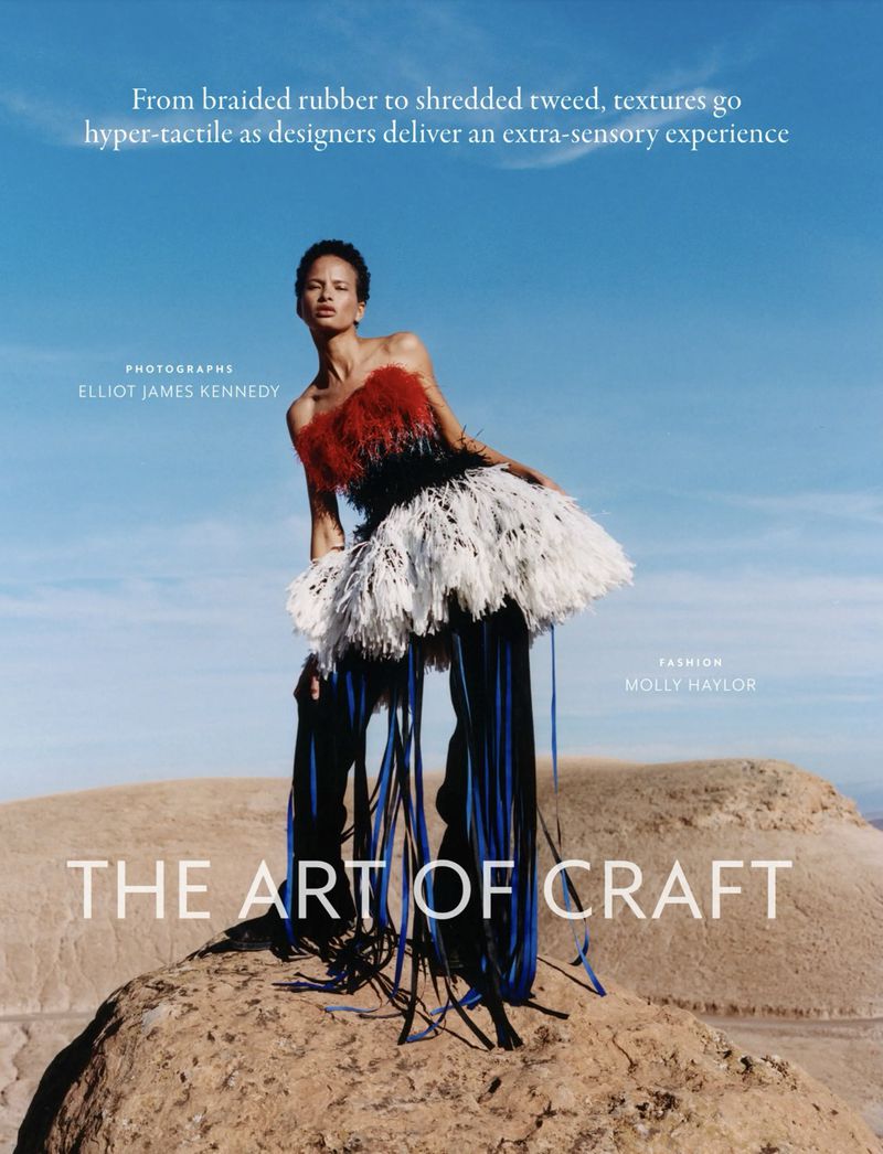 Fashion shot of an adult female model in a red, black, and white feather dress, posing on a rocky outcrop with a blue sky background.