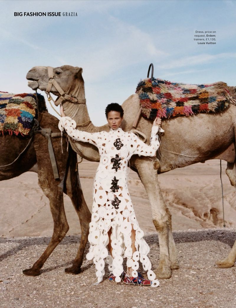 Fashion editorial shot of a Black female model in white dress posing with camels in desert landscape for Grazia Big Fashion Issue.