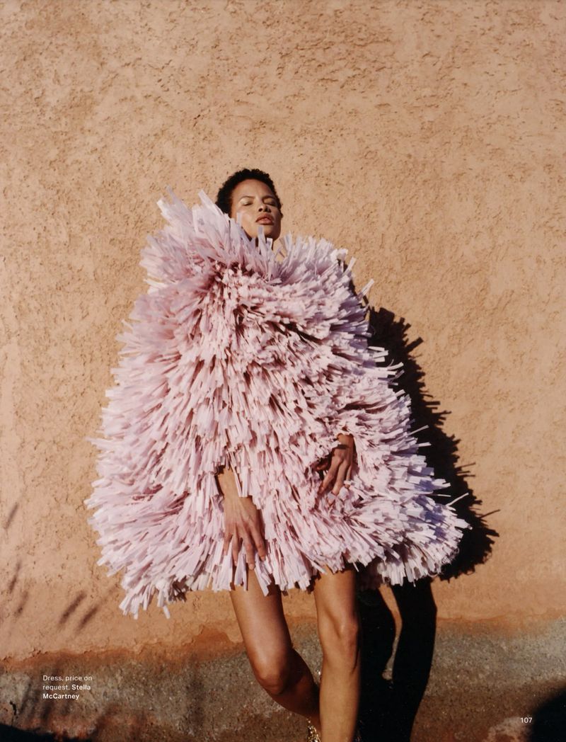 A woman poses outdoors, wearing a unique pink fringed dress. The shot captures a fashion-forward aesthetic with a textured wall backdrop.