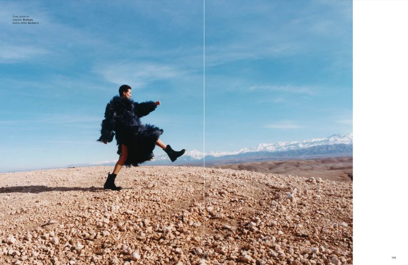 Fashion editorial photo featuring a woman in a furry coat and boots, walking on a rocky landscape with mountains and blue sky in the background.