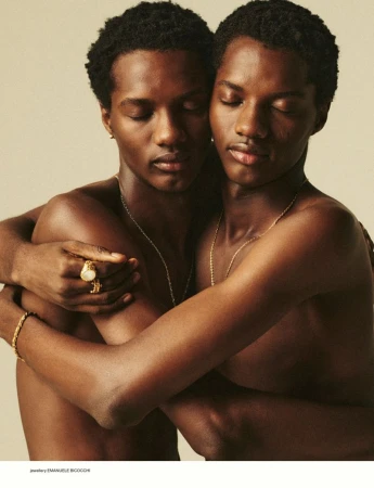 Close-up studio portrait of two dark-skinned adult males embracing, wearing minimal jewelry, against a neutral background.