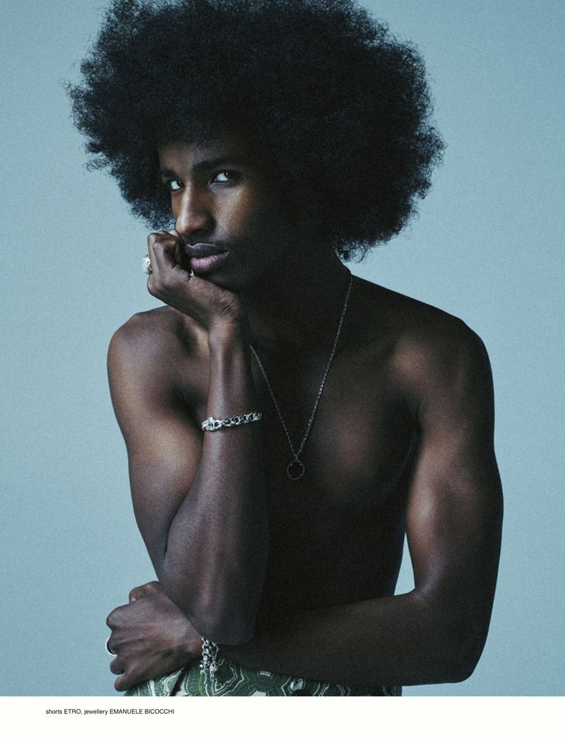 Close-up studio shot of a dark-skinned man with an afro, wearing a necklace and bracelet against a light blue background.