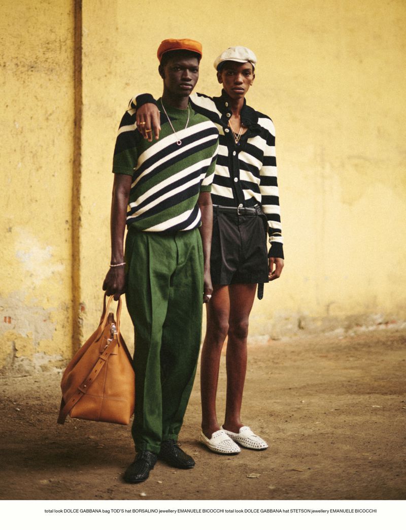 Two dark-skinned men pose in striped shirts, green pants, and shorts against a yellow wall. One carries a leather bag in a fashion shot.