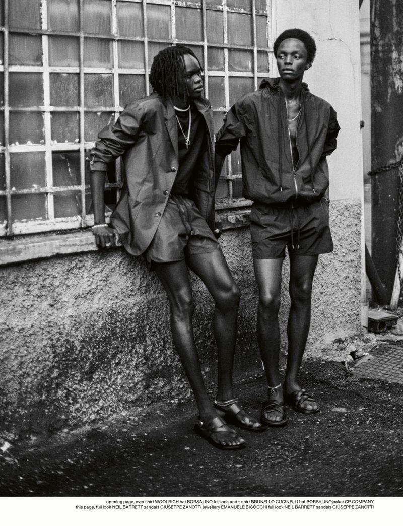 Monochrome fashion shot of two African men wearing shorts and jackets, standing against a windowed wall, evoking a cool, casual mood.