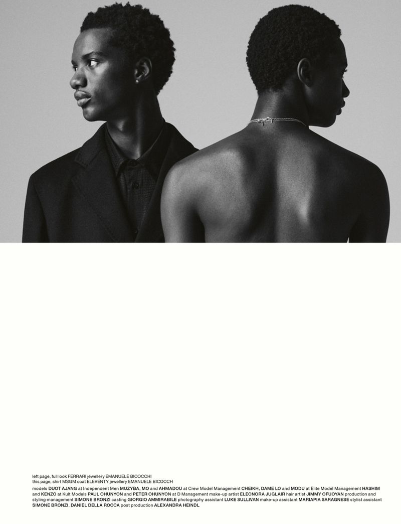 Two dark-skinned male models pose back to back, one wearing a black suit, the other shirtless with a silver chain. Black and white portrait.
